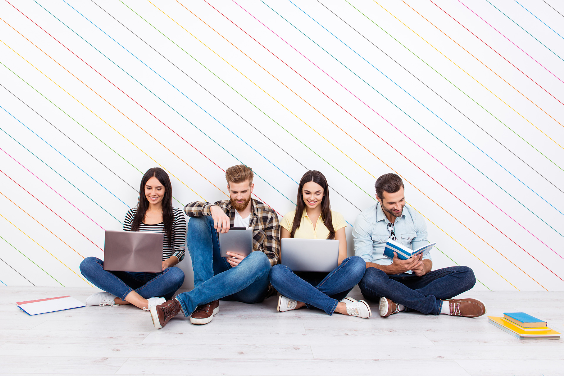 Group of friendly smiling students sitting on the floor and using modern technology for studying